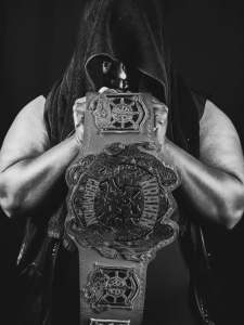 A Black professional wrestler named Brother Azriel wearing a black hood holds the Kraken Pro championship belt in front of his masked face.